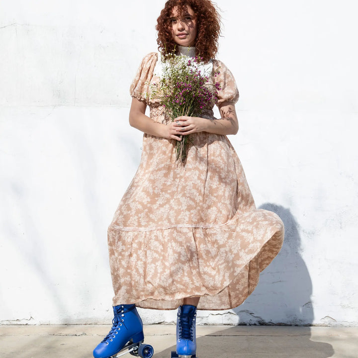 Woman in a floral dress with blue roller skates holding flowers against a white wall.
