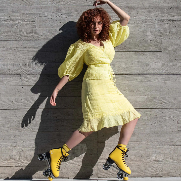 Woman in a yellow dress and roller skates posing against a wooden wall.