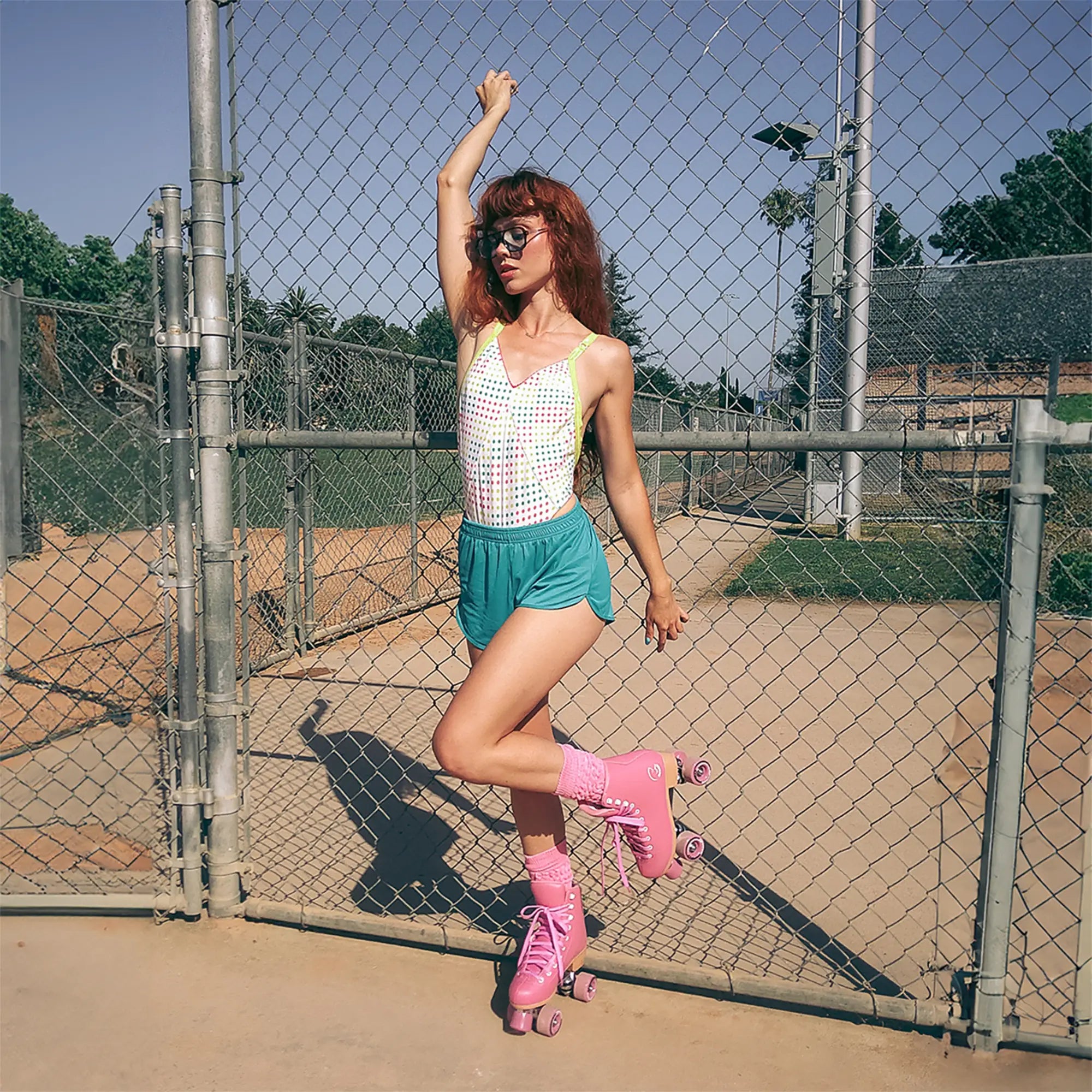 Woman in pink roller skates and green shorts standing in front of a chain-link fence.