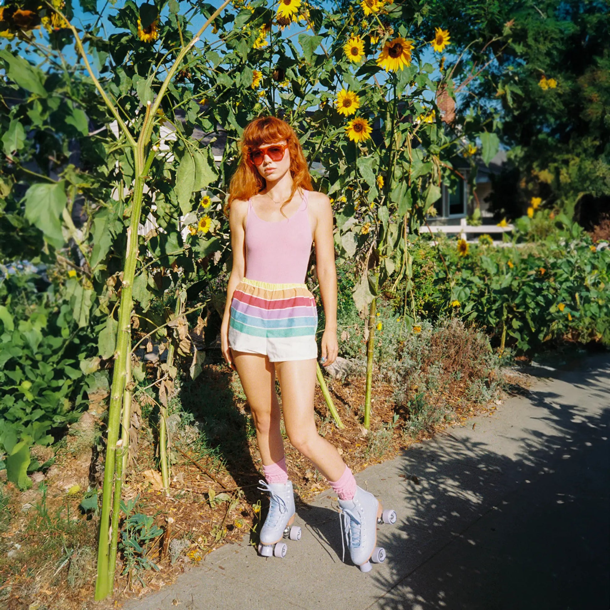 Person with red hair and sunglasses standing in a garden with sunflowers and greenery wearing roller skates