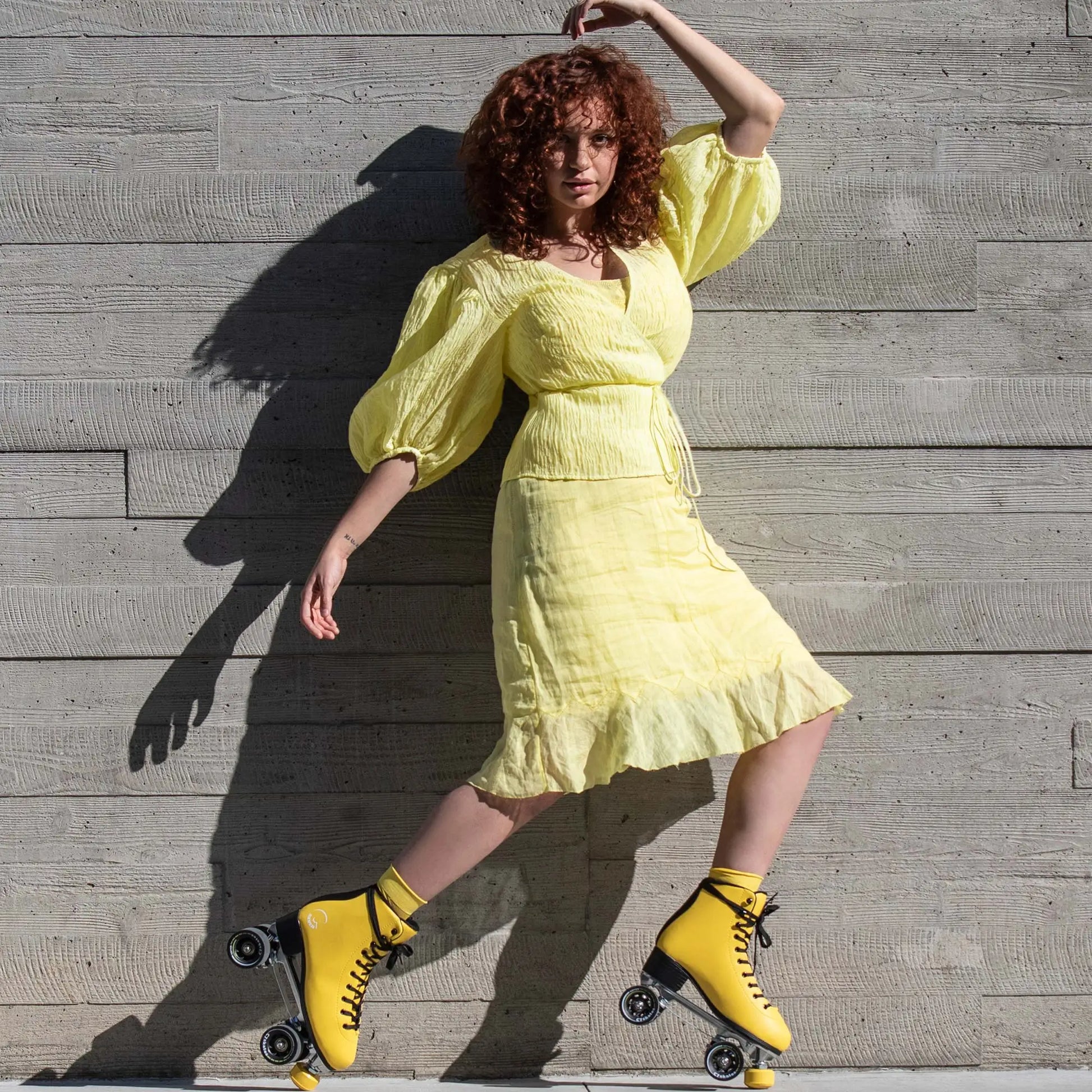 Woman in a yellow dress and roller skates posing against a wooden wall.