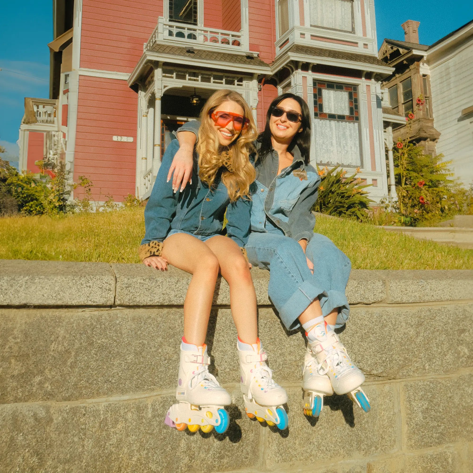 Two women sitting on steps in front of a house, wearing denim outfits and roller skates.