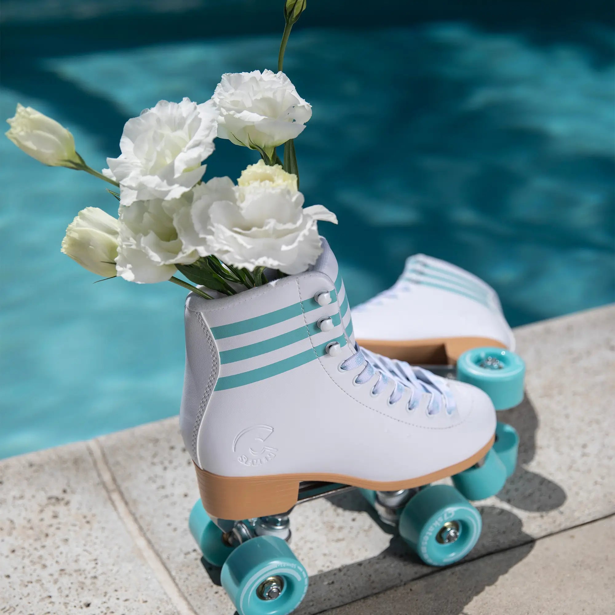 White roller skates with turquoise wheels holding white flowers by a pool.