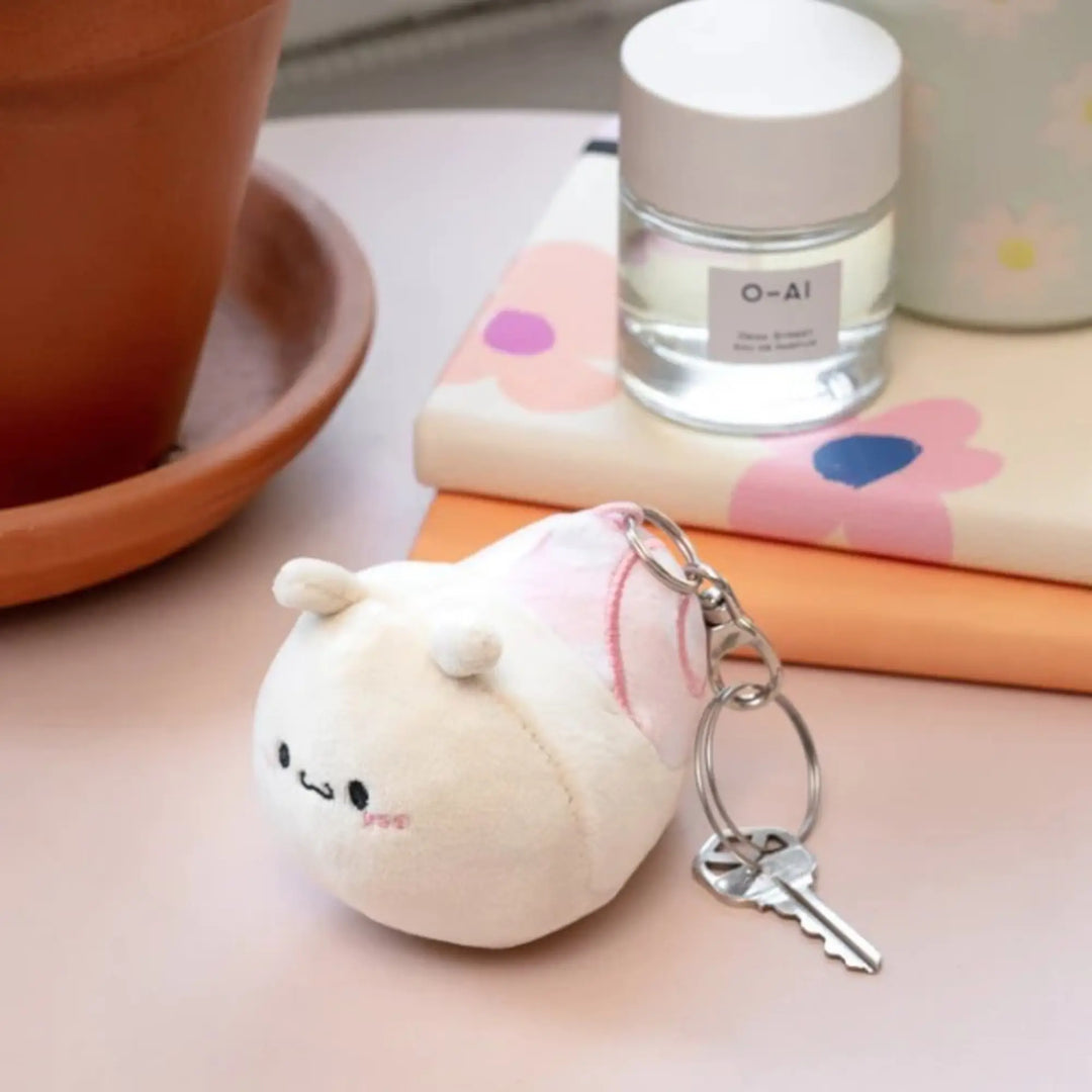 Plush keychain resembling a round creature with a key attached, placed on a surface with a bottle and books in the background.